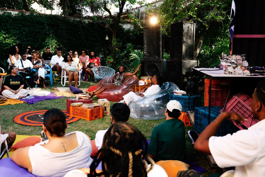 Roda de conversa num jardim, com o público sentado em almofadas, puffs e cadeiras